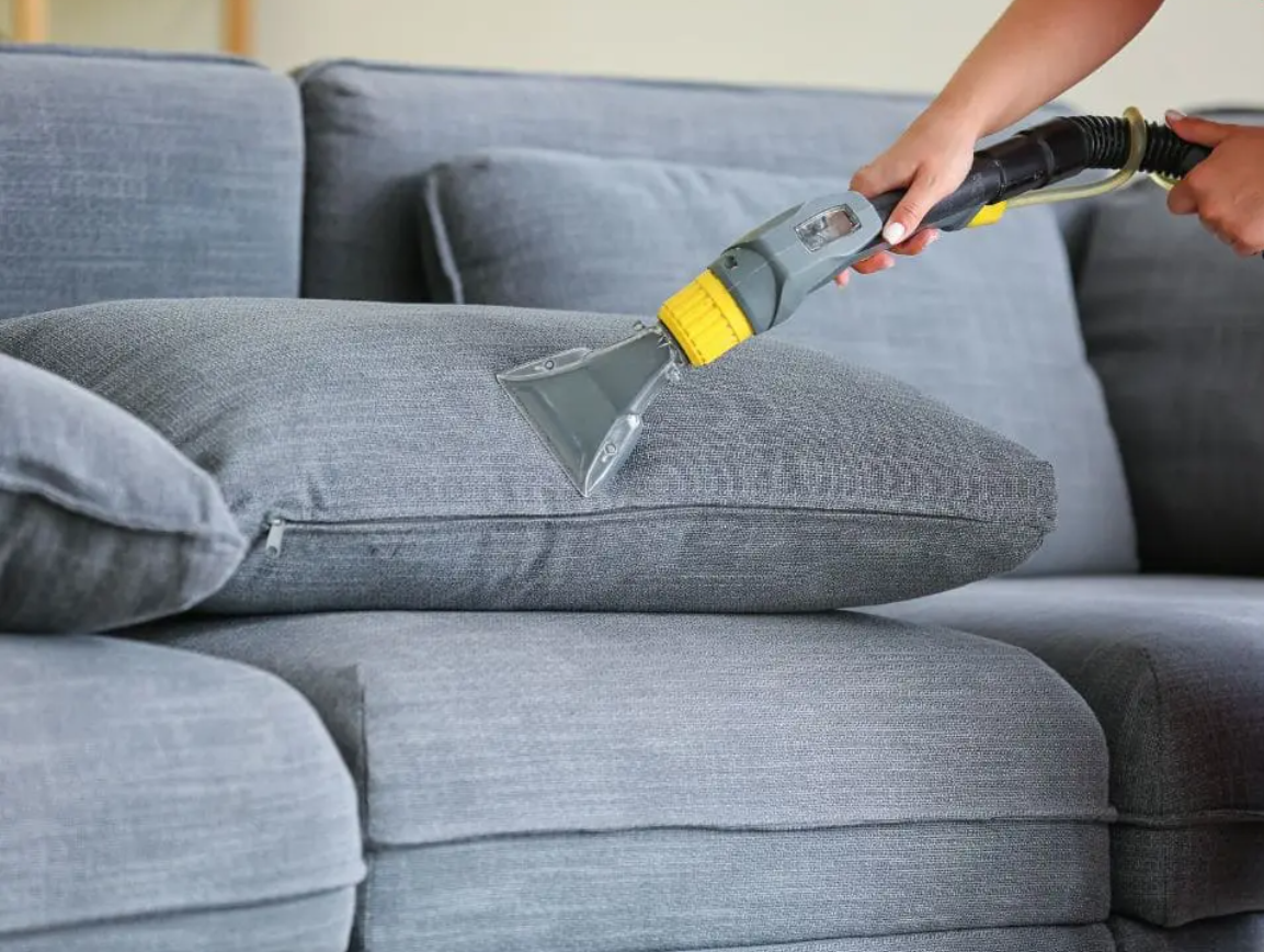 Osoba używająca odkurzacza do czyszczenia poduszki szarej sofy.
