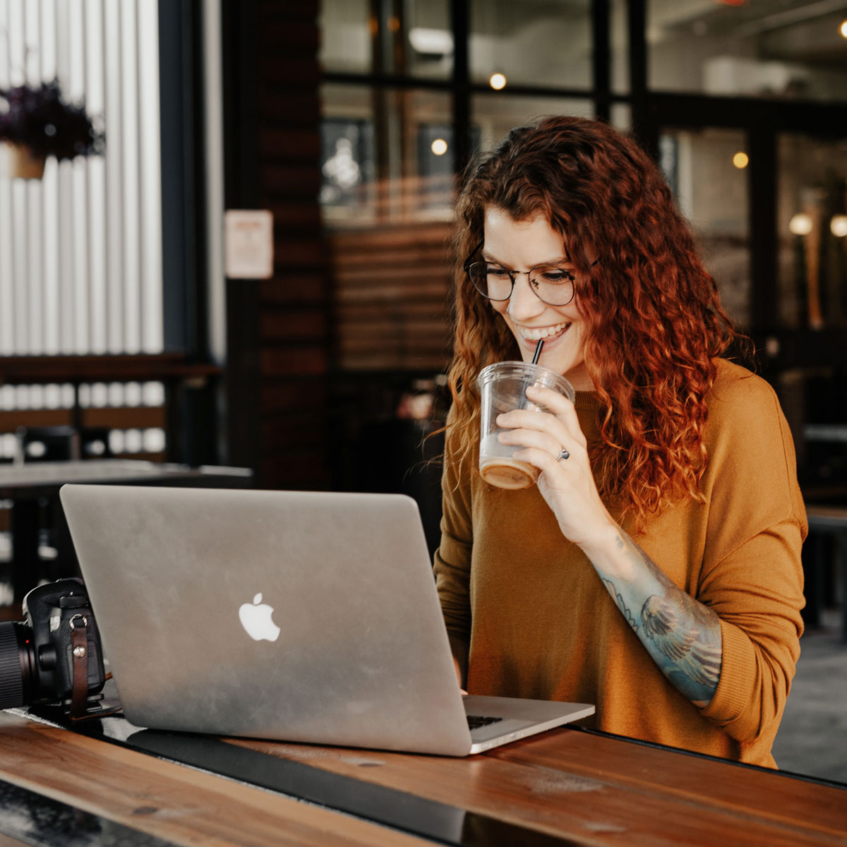 Kobieta z kręconymi włosami pije napój z plastikowego kubka, siedząc przed laptopem Apple.