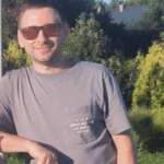 Mężczyzna w szarym t-shircie i okularach przeciwsłonecznych, stojący na tle zieleni.